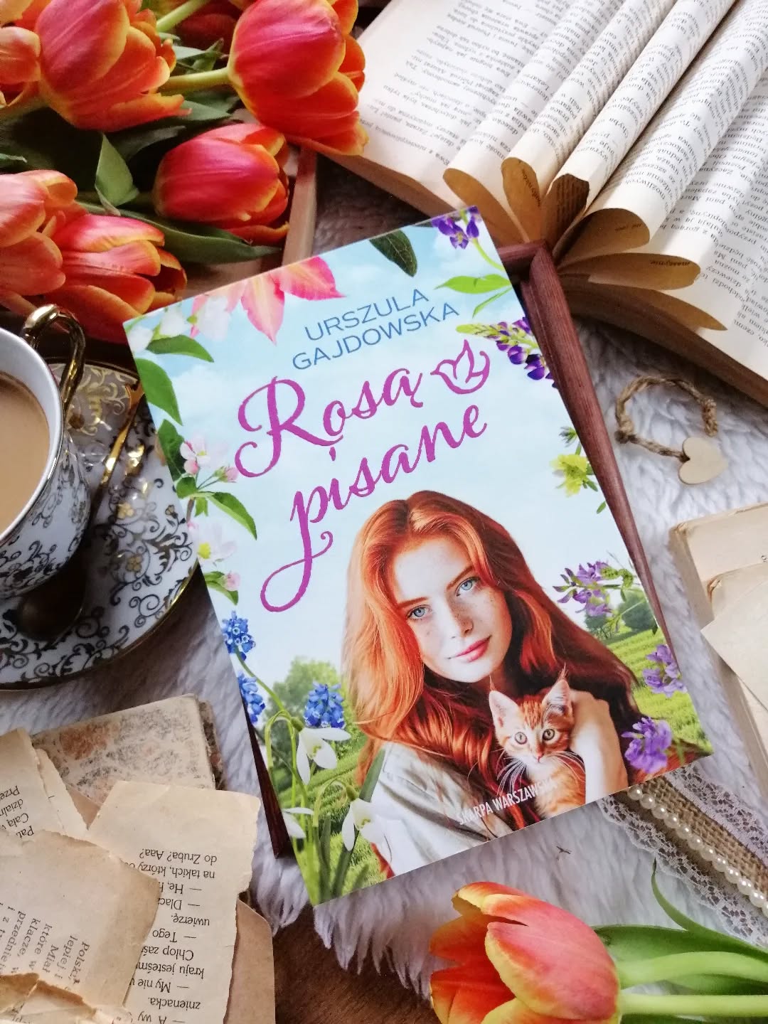 Dzień dobry 🤗  Na skraju lasu, w miejscu, które nazwano Azylem, zaczyna się historia o cichym powracaniu do życia.  “...najdotkliwsze uczucia rodzą się nie z tego, co powiedzieli, tylko z tego, czego nie odważyli się powiedzieć."  “Rosą pisane” Urszula Gajdowska  Edyta, po latach bólu i życia w cieniu, próbuje na nowo poukładać swoją codzienność. Razem z dziećmi Tosią i Tymkiem odnajduje coś, czego tak bardzo potrzebowała — spokój, bezpieczeństwo i oddech. Wśród ludzi, którzy również noszą w sobie swoje historie, i zwierząt, które leczą samą obecnością. Spokój Edyty zostaje zachwiany wraz z przyjazdem Dawida, byłego wojskowego. Mężczyzna niesie ze sobą ciężar doświadczeń z misji, które nauczyły go dystansu, a może nawet cynizmu…  Niby lekka, a jednak nie.  To historia subtelna, pełna niedopowiedzeń, a jednocześnie zwyczajna – z momentami ciszy i radościami jakie niesie codzienność. Autorka pięknie pokazuje dobre i złe strony życia, losy ludzi po przejściach – takich, którzy walczą o spokój, nie zgadzają się na zło i próbują stworzyć coś dobrego wokół siebie.  O zaufaniu, które wraca powoli. O ranach, które nie znikają od razu. O sile, która rodzi się gdzieś cicho — w zwykłych dniach, w małych gestach, w decyzji, żeby się nie poddać.  Bohaterowie są prawdziwi — nieidealni, z bagażem doświadczeń, z trudną przeszłością. Edyta, ciepła i silna. Dawid zamknięty w sobie, naznaczony tym, co widział i przeżył. I gdzieś pomiędzy nimi przestrzeń na coś, co nie przychodzi łatwo, ale może się wydarzyć…  Wciągająca, ciepła, życiowa, historia. Wywołuje wzruszenie i uśmiech. Przypomina, że nawet po najciemniejszych chwilach można znaleźć w sobie światło. Że warto walczyć o spokój, swoje szczęście. Bardzo polecam! Więcej link w bio.  Współpraca barterowa - @skarpawarszawska  #rosąpisane
powieść obyczajowa * emocje * książka * literatura kobieca * azyl